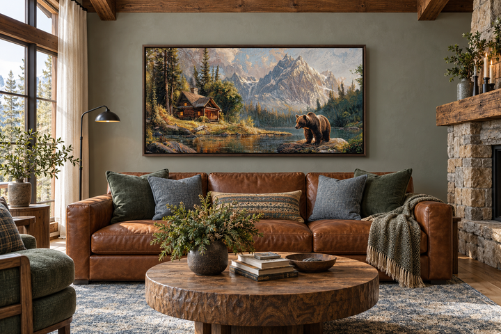 Framed panoramic canvas art of a bear near a mountain cabin above a brown leather sofa.