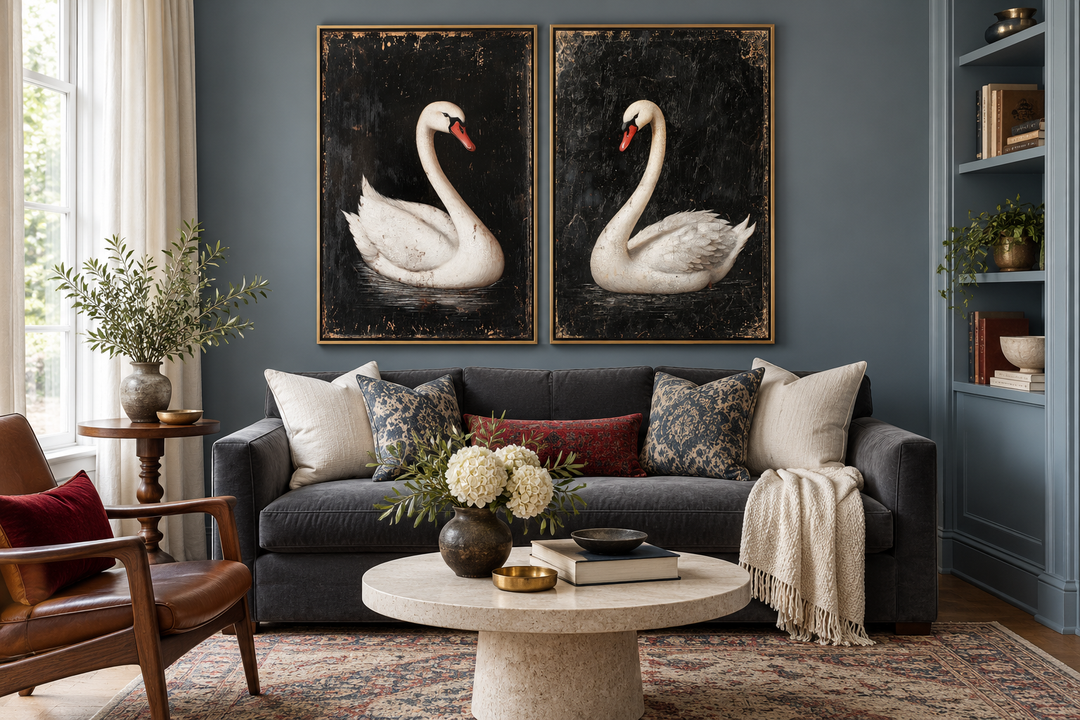 Set of 2 antique white swan paintings in gold frames on a dark blue wall above a grey sofa.