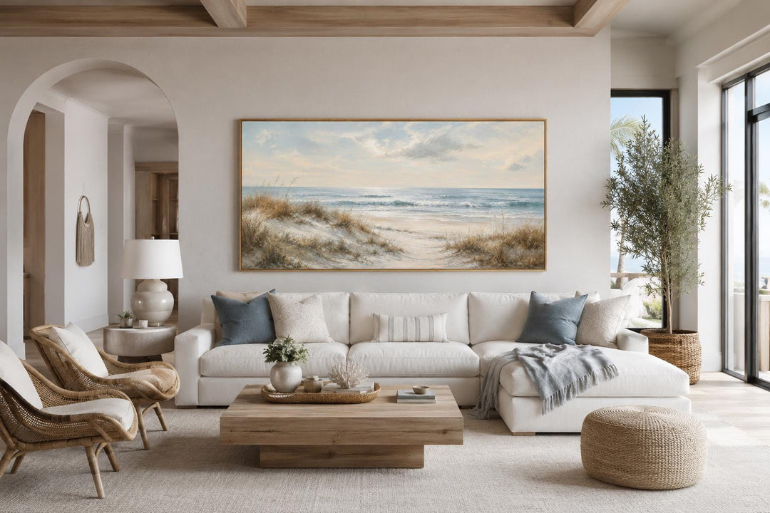 Sandy beach dunes wall art with sea grass and rolling waves in a slim gold frame above a white sofa.