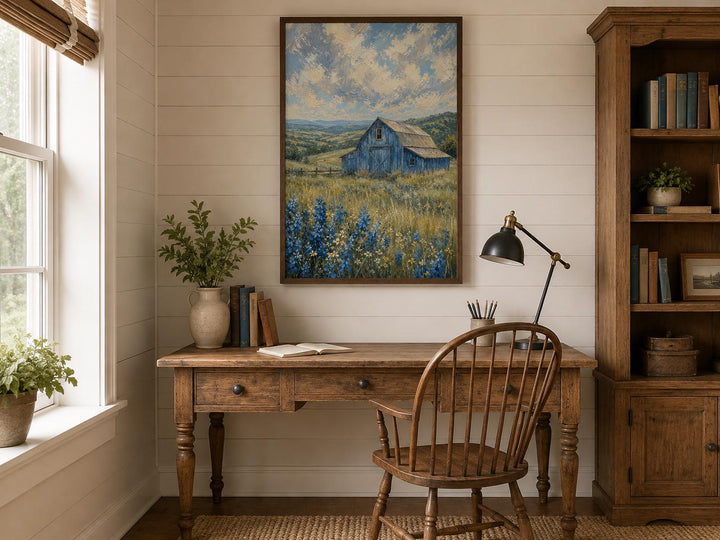 Vertical canvas art of a blue barn in a field of bluebonnets hanging above a rustic wooden desk.