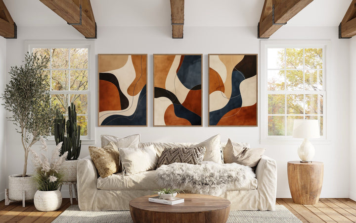 3 piece mid-century modern abstract wall art in earth tones, brown, cream, and navy above a sofa.