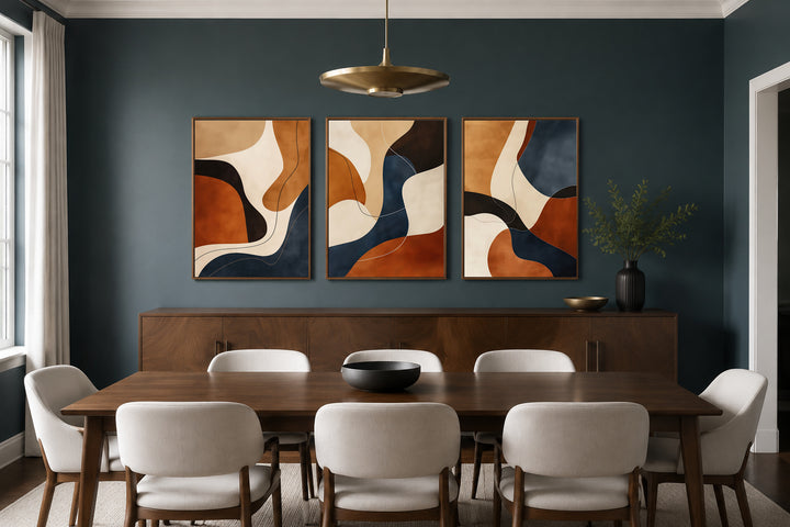 3 piece mid-century modern abstract wall art in earth tones, cream, and navy above a dining table.