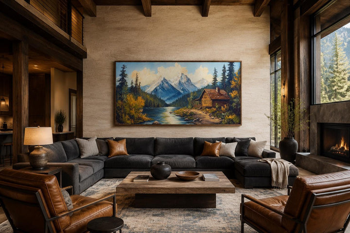Vintage style canvas wall art of a mountain cabin by a river, hung above a large sectional sofa.