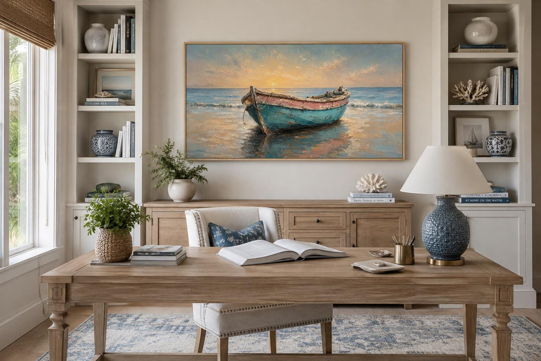 Weathered teal and pink rowboat painting at sunset in a gold frame above a wooden office desk.