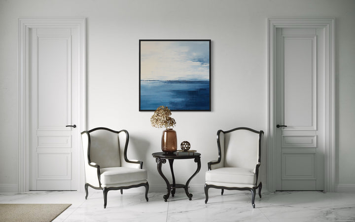 Abstract ocean square framed canvas wall art in navy blue, grey, and silver above two white chairs.