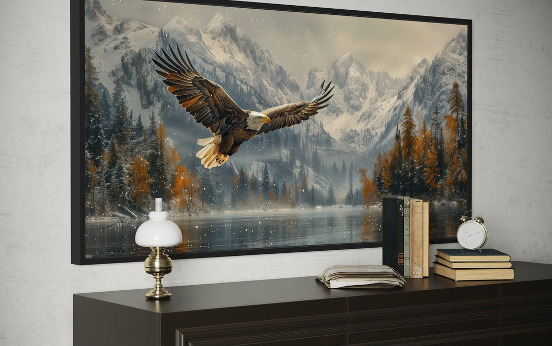 Framed wall art of a bald eagle flying over a snowy mountain lake above a dark wood dresser.
