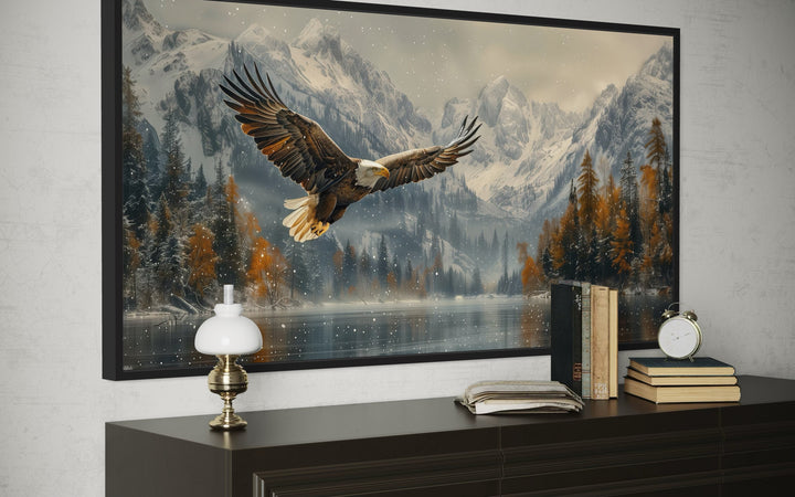 Framed wall art of a bald eagle flying over a snowy mountain lake above a dark wood dresser.