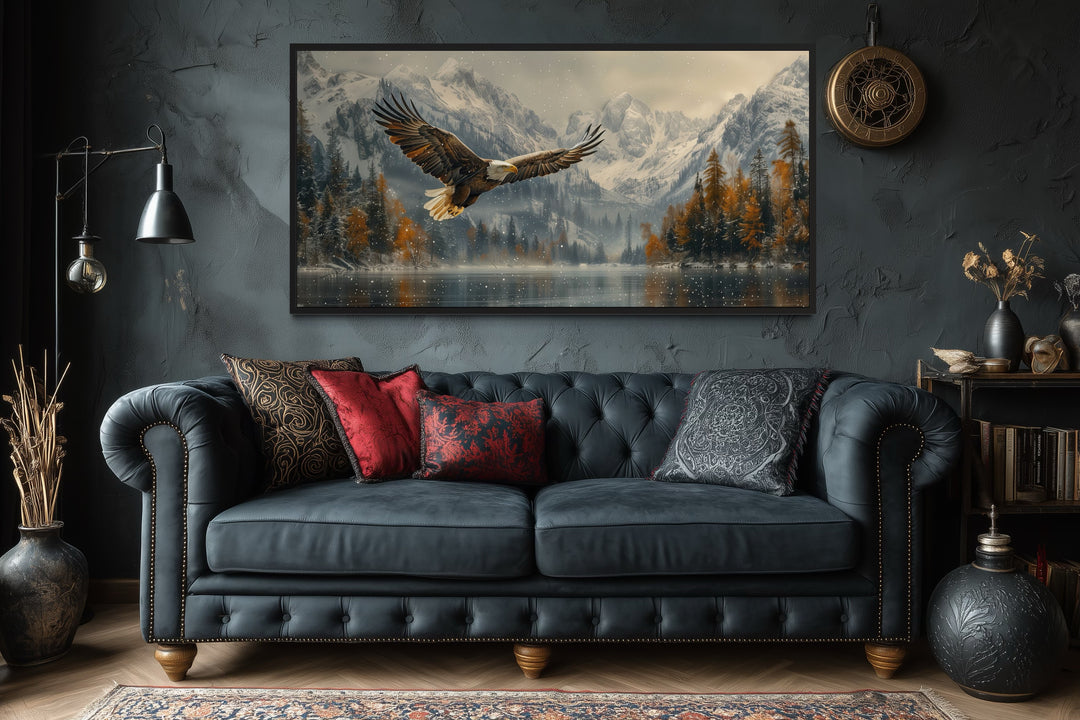 Framed wall art of a bald eagle flying over a snowy mountain lake above a dark tufted sofa.