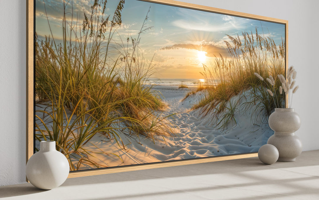 Framed canvas wall art of a beach sunset with sand dunes and sea grass in a coastal room setting.