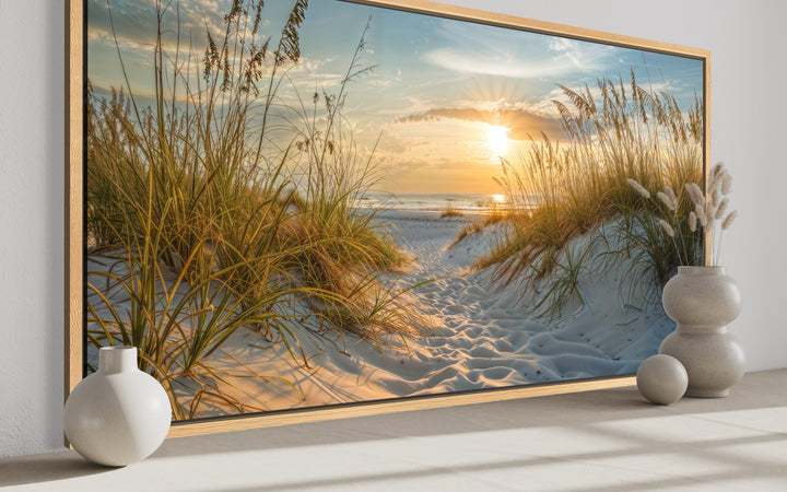 Framed canvas wall art of a beach sunset with sand dunes and sea grass in a coastal room setting.