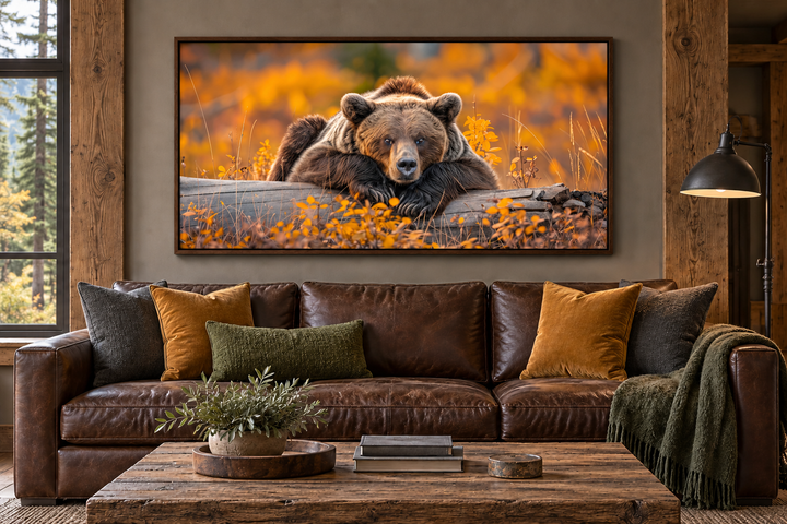 A framed canvas print of a grizzly bear resting on a log in an autumn forest hangs above a sofa.
