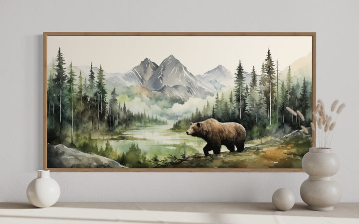 Framed watercolor painting of a bear in a mountain forest, displayed above a minimalist shelf.