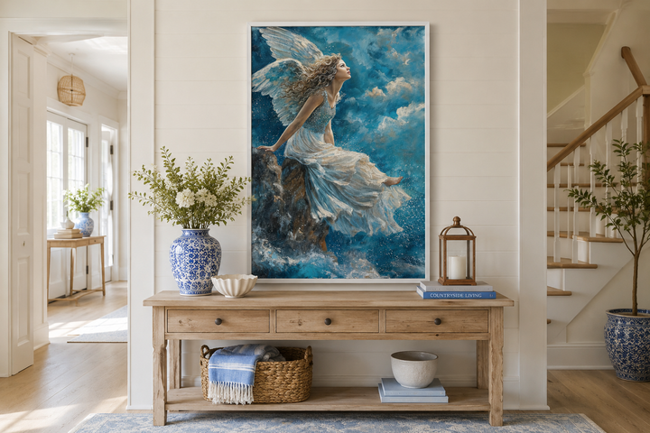 Framed wall art of an angel in a white dress sitting on a mountain peak among blue clouds.