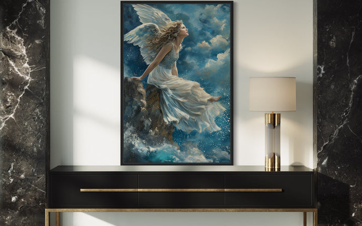 Framed wall art of a winged angel in a white dress sitting on a rock above crashing blue waves.