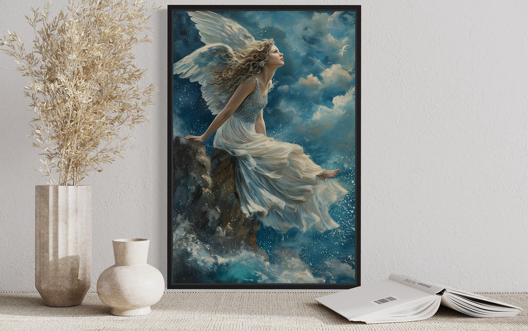 Framed wall art of a winged angel in a white dress sitting on a mountain peak among blue clouds.