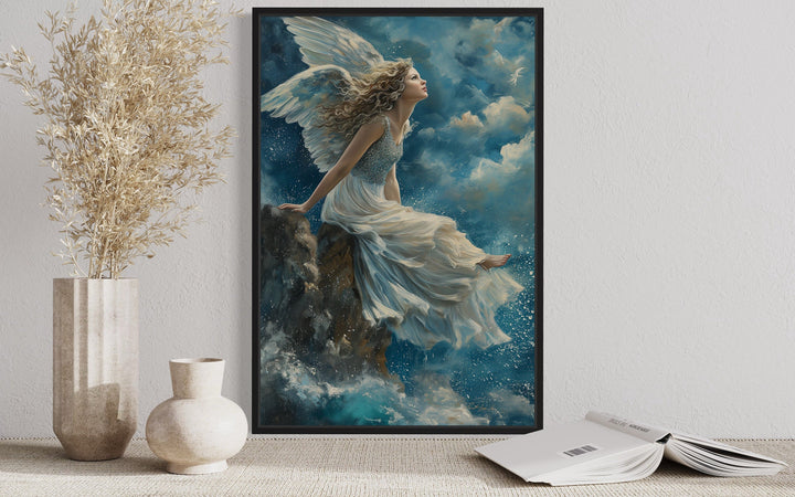 Framed wall art of a winged angel in a white dress sitting on a mountain peak among blue clouds.