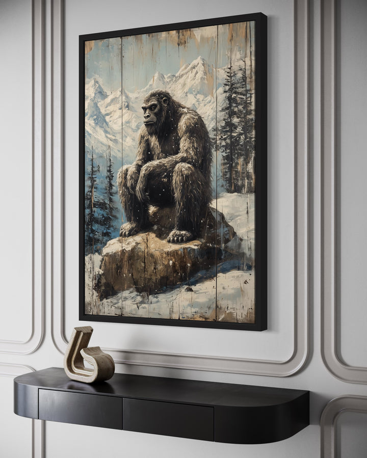 Framed canvas wall art of Bigfoot sitting on a rock in a snowy mountain landscape with pine trees.