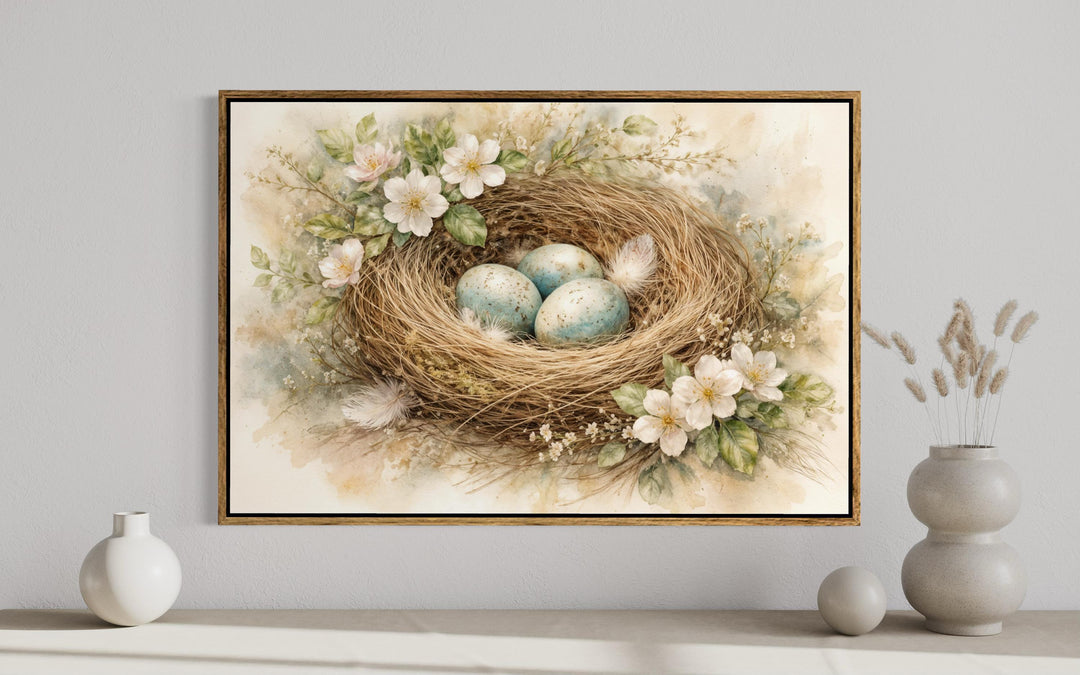 A framed watercolor canvas print of a bird's nest with blue eggs and spring flowers on a wall.