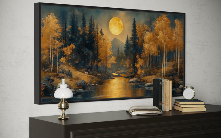 Framed canvas wall art of a moonlit forest with golden trees and a river on a dark dresser.