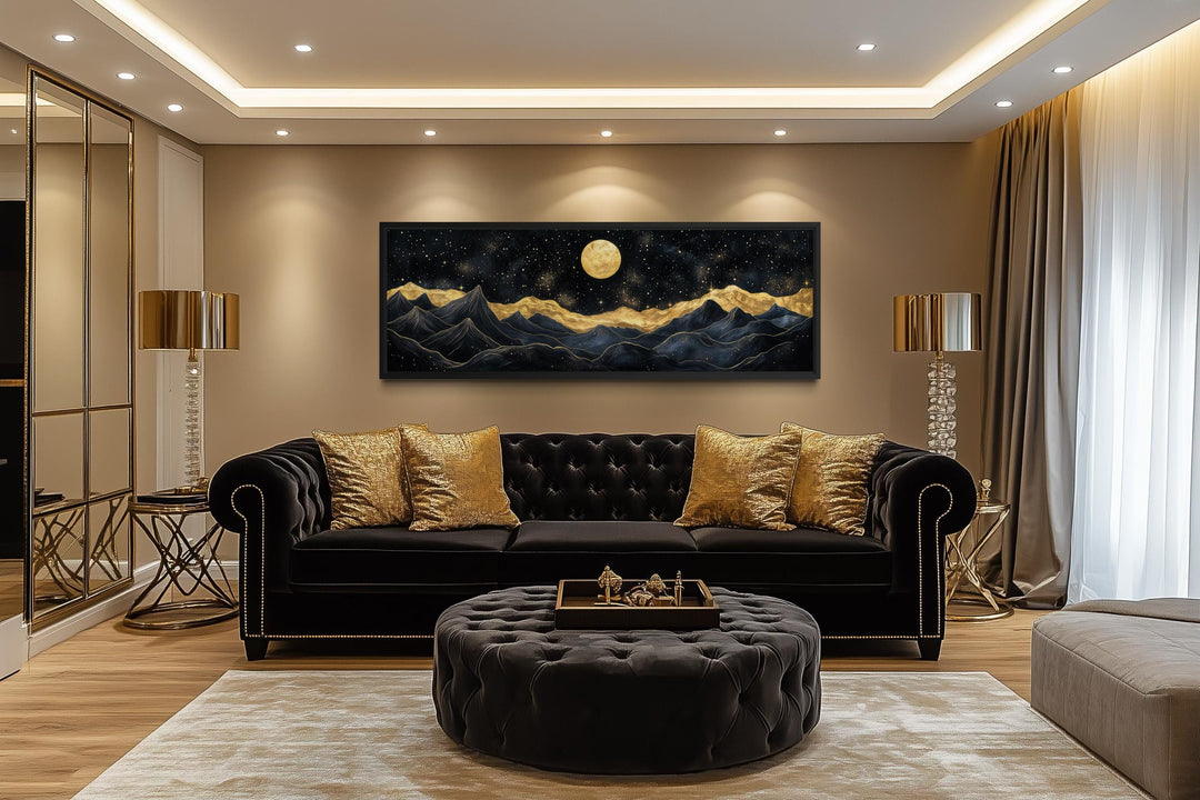 A panoramic canvas print of a moonlit mountain landscape in black and gold hangs above a black sofa.