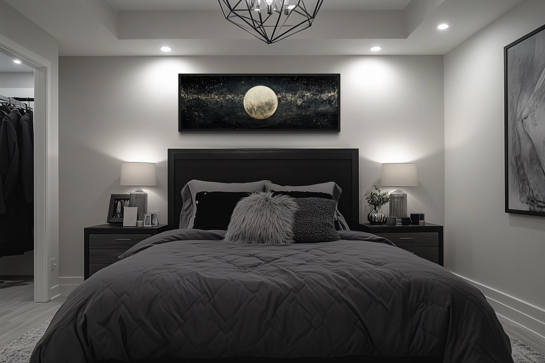 Horizontal black and white canvas art of a full moon in a starry sky hanging above a bed.