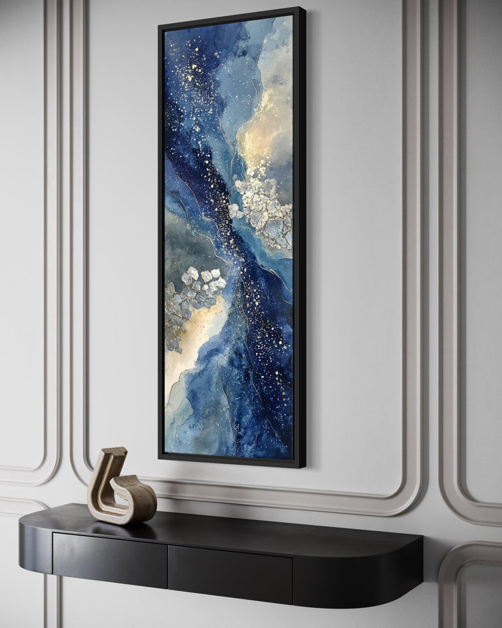Tall narrow abstract canvas print with navy, beige, and gold tones in a black frame on a gray wall.