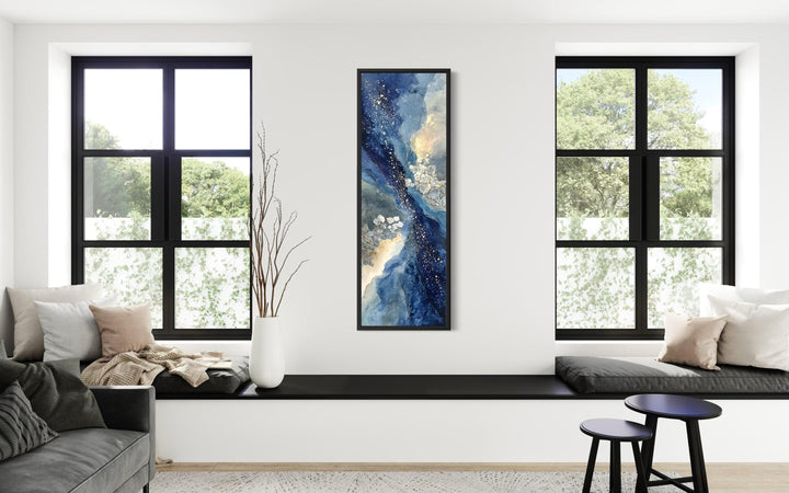 Tall narrow abstract canvas print with blue, beige, and gold tones on a white wall between windows.
