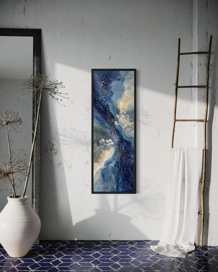 Tall narrow abstract canvas print with navy, beige, and gold tones in a black frame on a white wall.