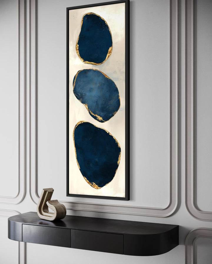 Vertical narrow canvas print featuring three abstract blue stones with gold edges on a light wall.