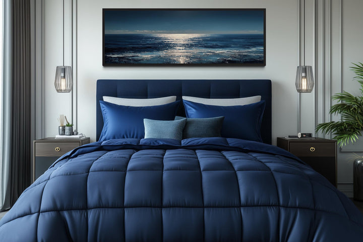 A long horizontal seascape painting of a moonlit blue ocean hangs above a modern navy blue bed.