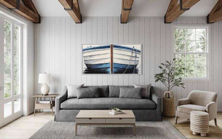 Blue and white rustic boat painting canvas wall art centered above a grey sofa in a coastal room.