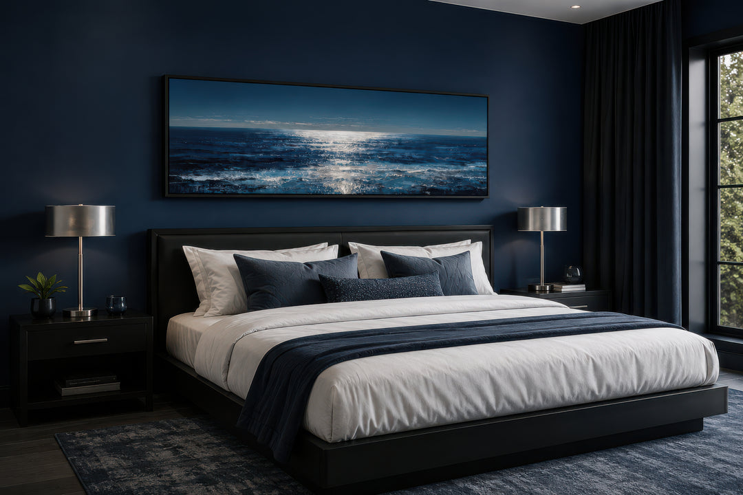 A long horizontal canvas of a moonlit blue and silver ocean seascape hangs above a modern bed.