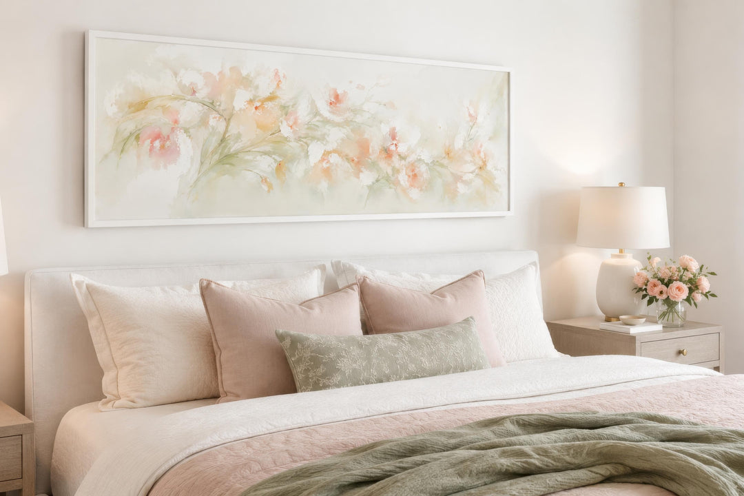 Blush pink floral watercolor canvas painting in a white frame hanging above a bed.