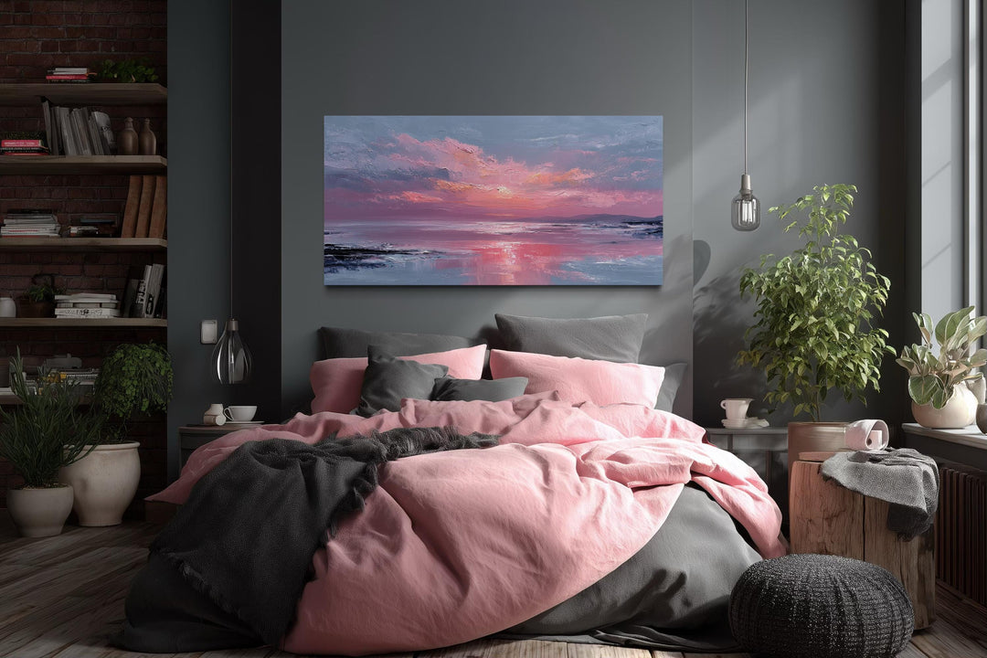 A horizontal canvas print of a pink ocean sunset hangs above a bed with pink and grey linens.