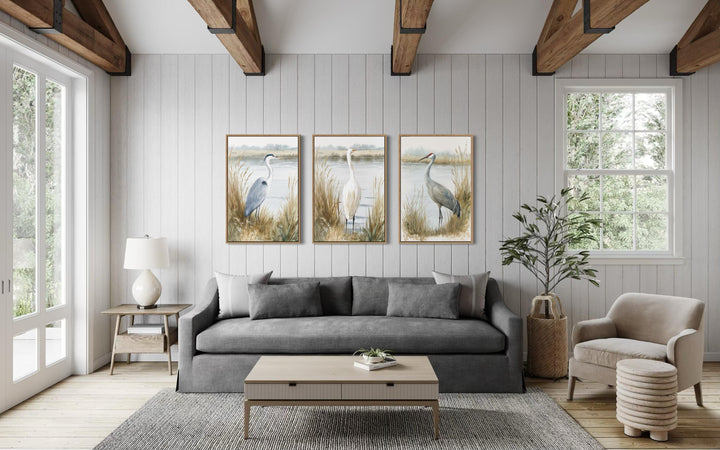 A triptych of framed coastal bird prints featuring a heron, egret, and crane over a gray sofa.