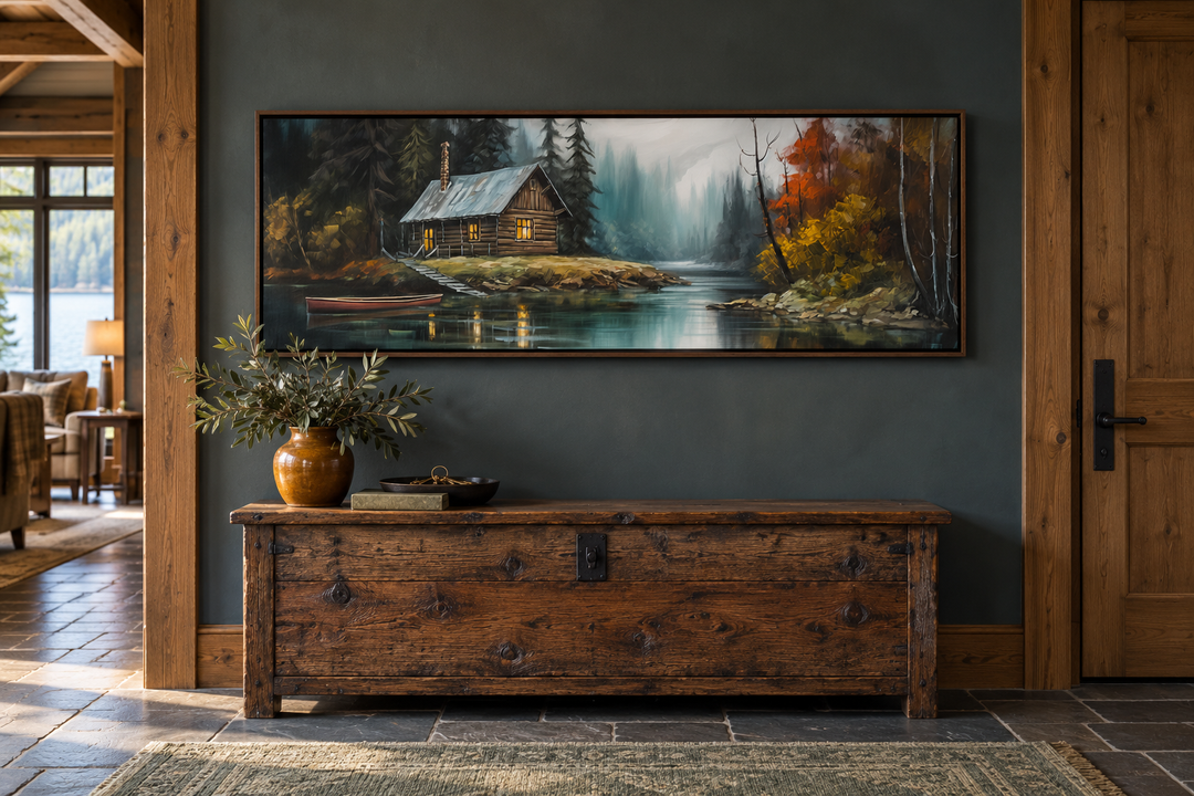 Narrow rustic framed canvas wall art of a cabin on a lake above a wooden chest in an entryway.