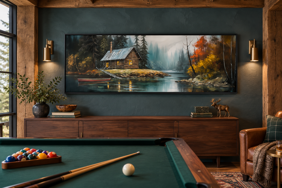 Narrow horizontal framed canvas art of a rustic cabin on a lake above a wooden sideboard.