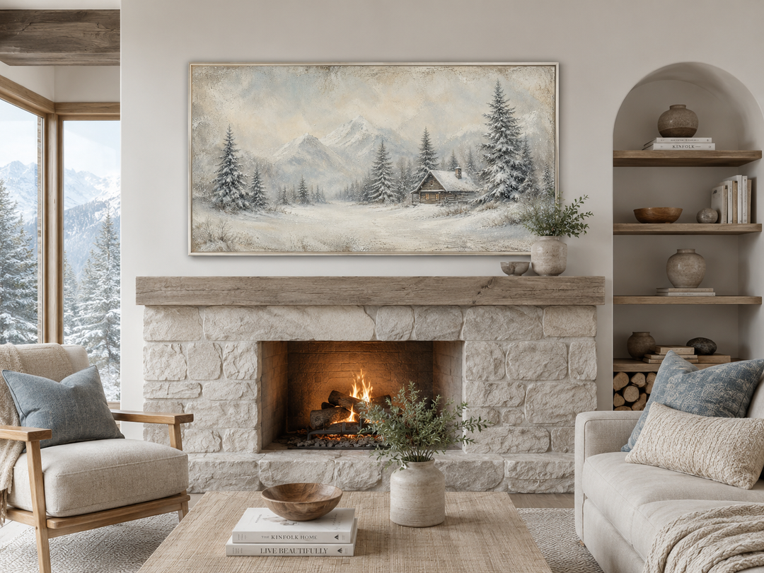 Rustic framed canvas art of a snowy mountain cabin hanging above a stone fireplace in a cozy room.
