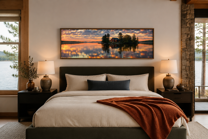 Long horizontal framed canvas wall art of a cabin on a lake at sunset above a bed.