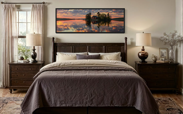 Long horizontal framed canvas wall art of a cabin on a lake at sunset above a dark wood bed.