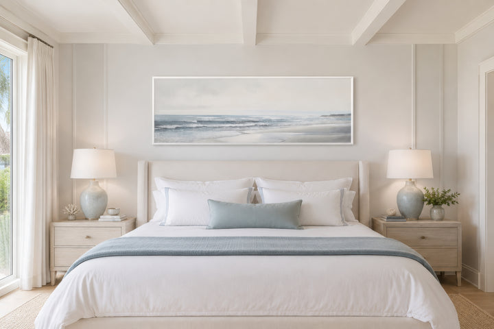 A long horizontal framed canvas featuring a minimalist coastal landscape above a neutral bed.
