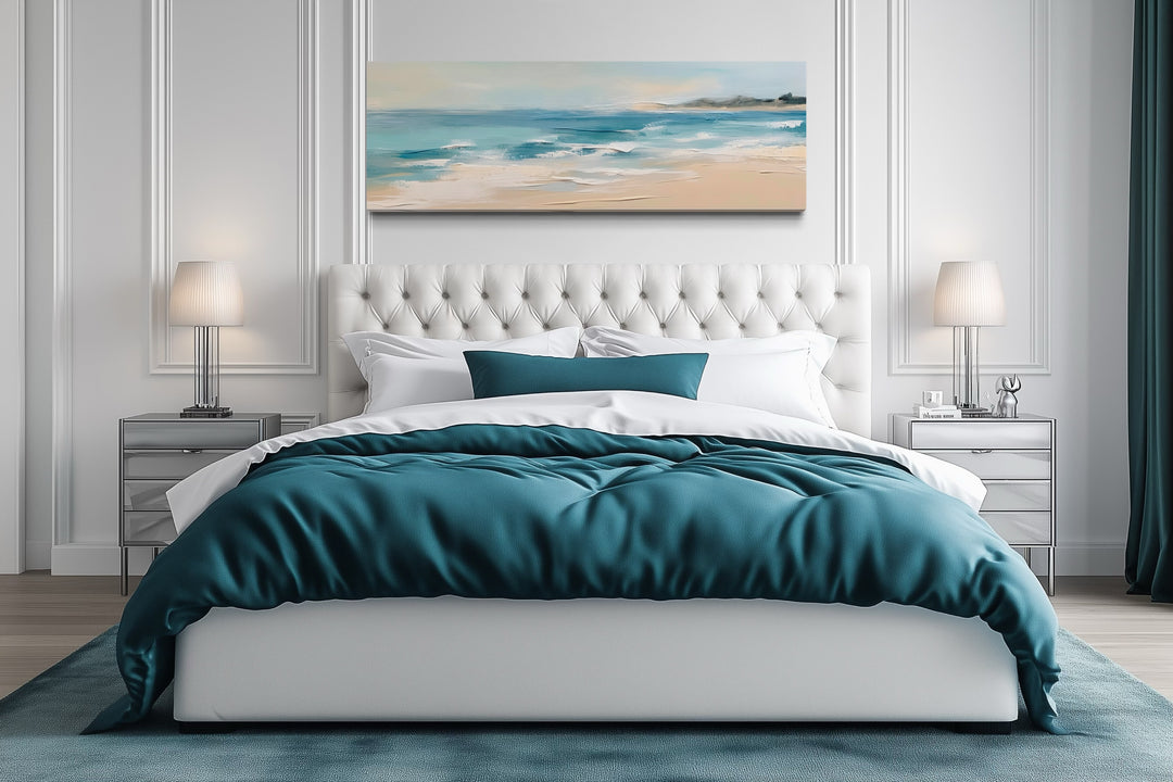 Panoramic coastal canvas wall art of a calm ocean beach hanging over a tufted white bed.