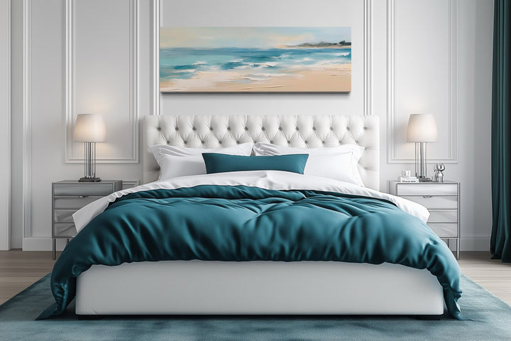 Panoramic coastal canvas wall art of a calm ocean beach hanging over a tufted white bed.