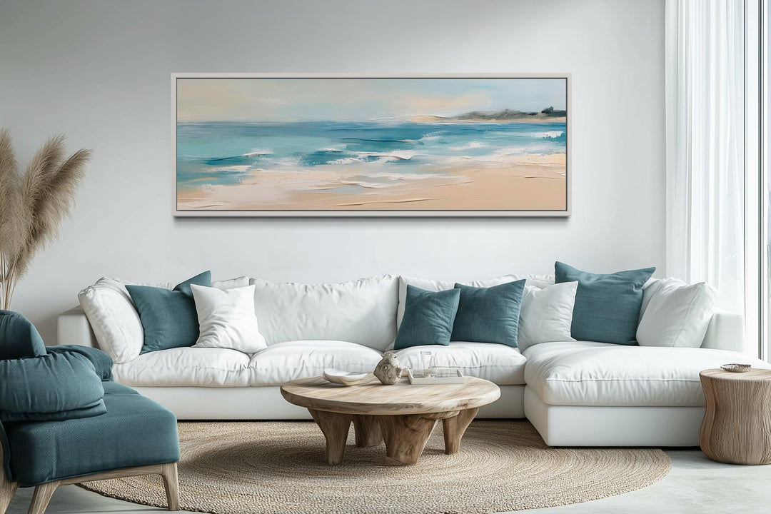 Panoramic coastal framed canvas wall art of a calm ocean beach above a white sectional sofa.