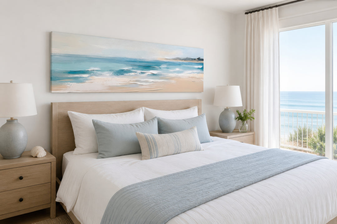 Panoramic coastal canvas wall art of a calm ocean beach hanging over a bed in a neutral bedroom.