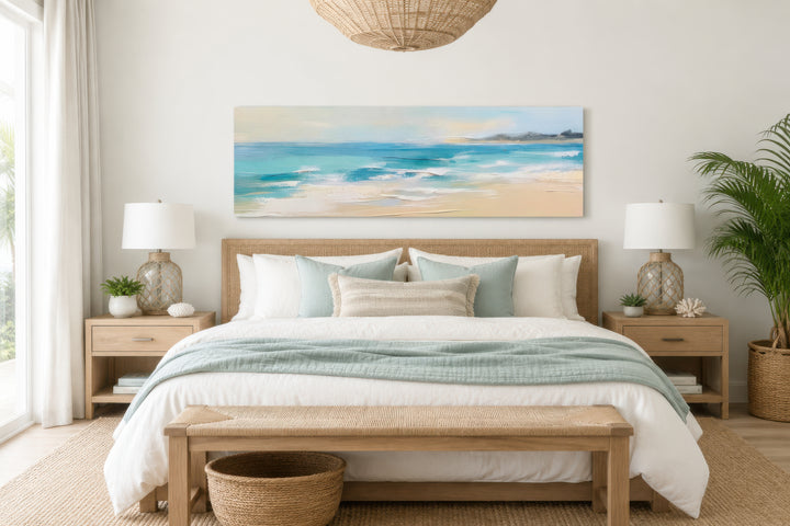 Panoramic coastal canvas wall art of a calm ocean beach hanging over a bed in a neutral bedroom.