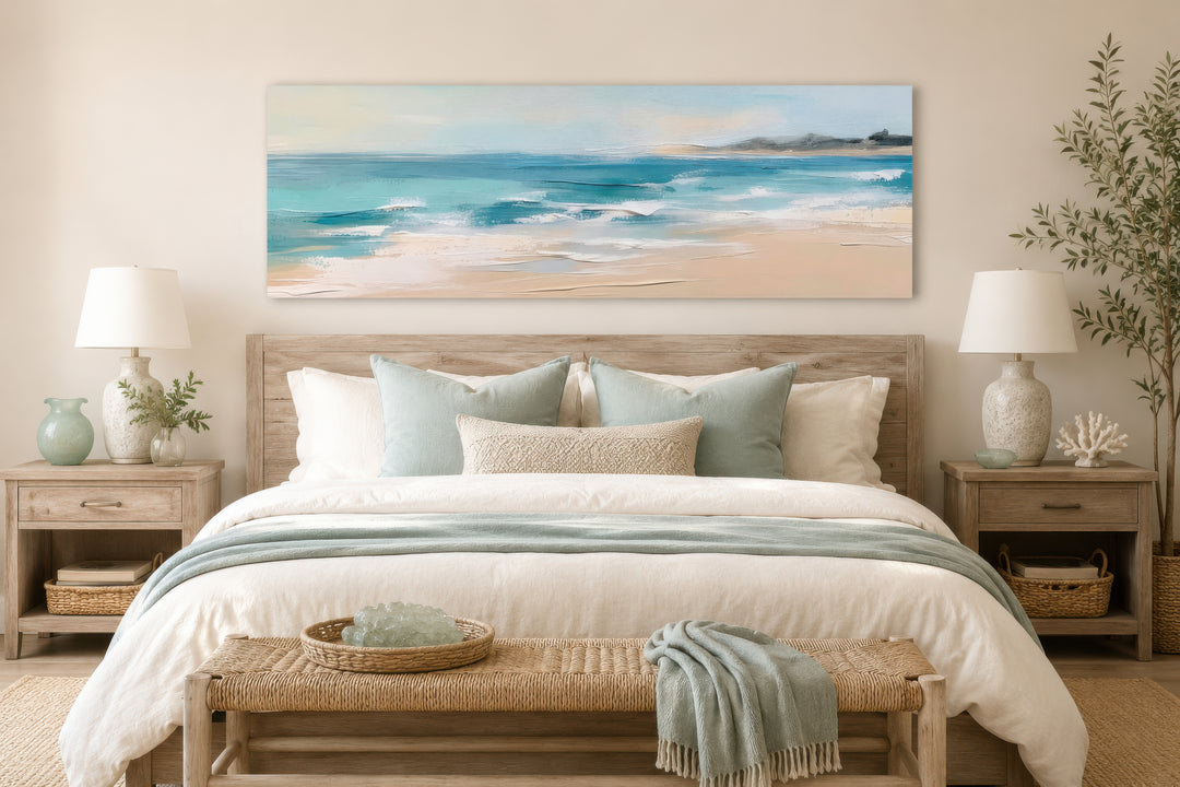 Panoramic coastal canvas wall art of a calm ocean beach hanging over a bed in a neutral bedroom.