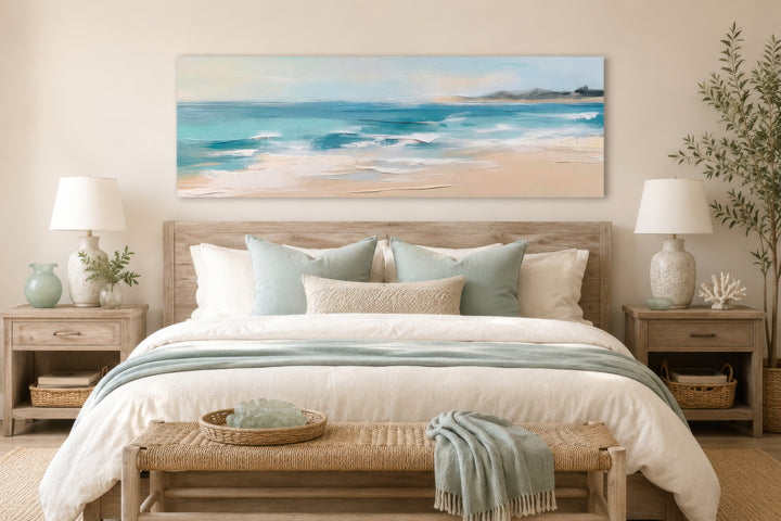 Panoramic coastal canvas wall art of a calm ocean beach hanging over a bed in a neutral bedroom.