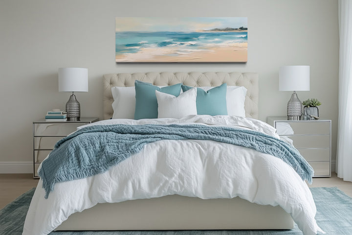 Panoramic coastal canvas wall art of a calm ocean beach hanging over a bed in a modern bedroom.