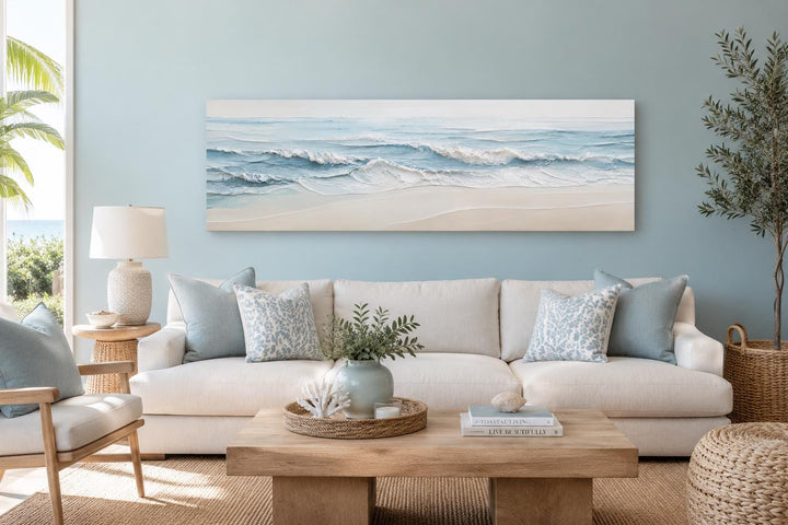 Wide horizontal canvas of calm ocean waves in soft blue and cream tones above a white sofa.